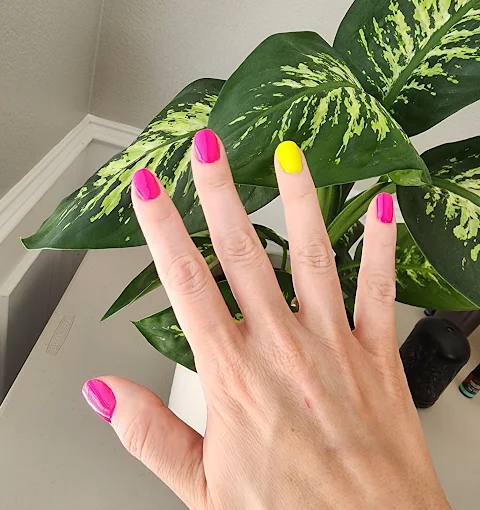 Bright neon pink and yellow manicure beside a leafy plant